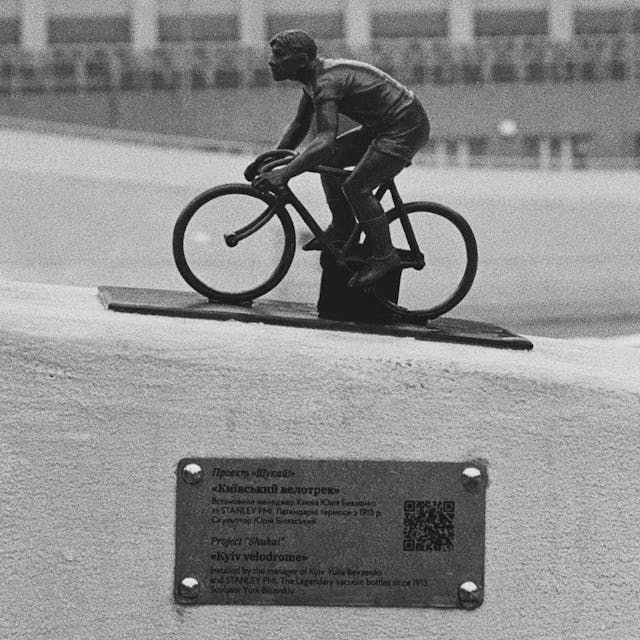 A statue of a biker. The statue has a label below that describes it and uses a QR code to let people read more about it on the internet. Picture is in grayscale.