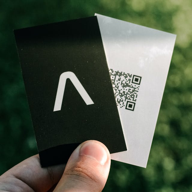 A hand holding business card that has a QR code printed on it. Grass in the background