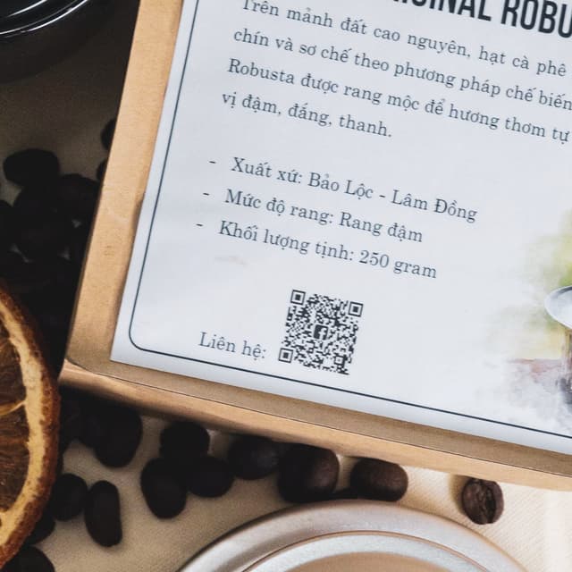 A bag of coffee is laying on a table near a cup of coffee. The bag has a QR code printed on the front. The code is used to let people go to the product's website. There are orange slices on the bottom left corner, and coffee beans in the background.